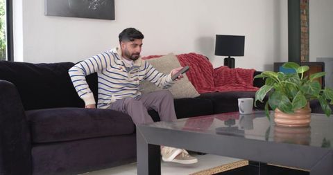 Indian man relaxing on living room sofa holding remote control, cozy modern home interior