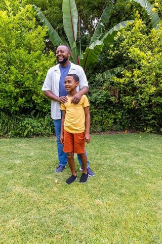 African american father and son smiling in lush garden, father resting hands on son's shoulders