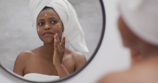 Image shows woman with towel on head applying cream to face while looking at bathroom mirror. Represents concepts of self-care, beauty routine, daily hygiene, and personal care. Ideal for marketing skincare products, wellness blogs, health articles, and beauty tutorials.