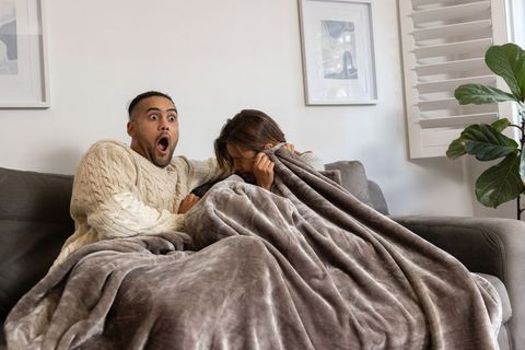 Couple reacting to scary movie while cuddling under plush blanket on couch