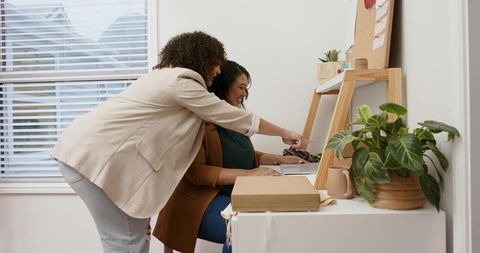 Women Collaborating at Home Office Desk Pointing at Laptop Screen with Plant and Corkboard
