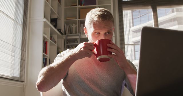 Happy caucasian man drinking from cup and looking on laptop in sunny living room. Lifestyle, wellbeing, relaxation, free time and domestic life, communication, unaltered.