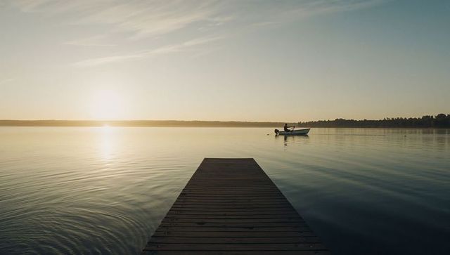 Wooden dock leading into calm lake at golden hour during sunrise with solo motorboat silhouette