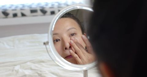 Asian woman checking under-eye skin in tabletop mirror on bed during daytime self-care