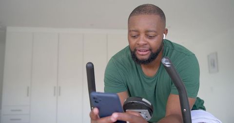 African man checking smartphone while leaning on stationary bike at home