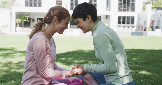 Two women are outside on a grassy lawn, smiling and holding hands. Both are dressed in casual clothing suitable for outdoor activities. This image can be used to depict themes of friendship, bonding, happiness, connection, and positive relationships. Ideal for websites and publications focused on lifestyle, wellness, outdoor activities, and personal connections.