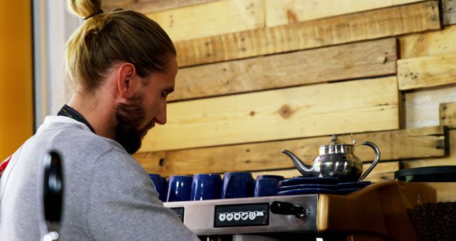 Young barista with man bun brewing coffee in a rustic cafe with wooden interiors. Ideal for depicting the modern cafe culture, coffee shops, or young professionals at work. Suitable for articles or advertisements about coffee making, cafes, barista training, or lifestyle blogs.