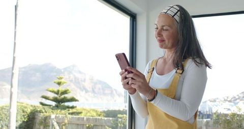 Woman texting on phone in sunlit coastal home with terrace garden and mountain view