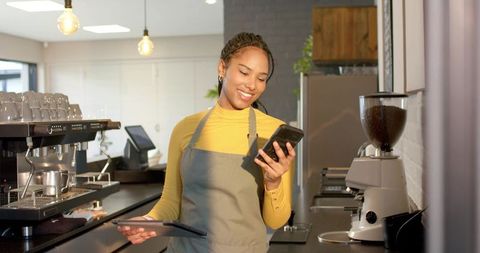 African american barista managing orders using smartphone and tablet at modern coffee counter