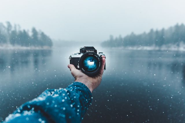 Person holds vintage Nikon camera over river surrounded by winter landscape. Snowfall creates serene atmosphere. Perfect for themes of nature photography, winter aesthetics, peacefulness, and outdoor exploration.