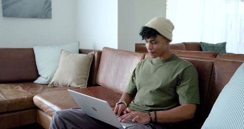 African man working remotely on laptop from modern leather sofa in bright living room