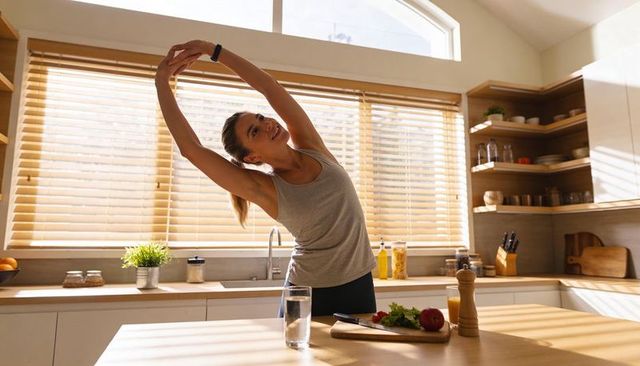 Woman stretching in sunlit kitchen morning routine with healthy snack and water