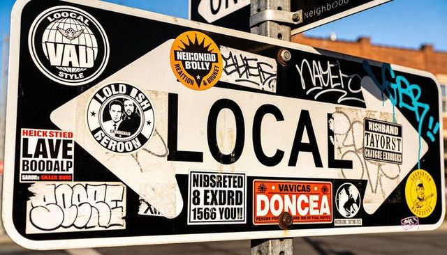 Sticker-covered LOCAL sign pointing right on urban pole with graffiti and rusted bolt
