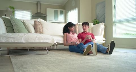 Diverse couple sitting on living room floor with mugs in modern bright cozy home
