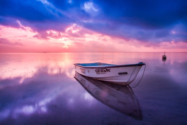 This stunning scene captures a serene sunrise over a calm and tranquil sea with an anchored boat reflecting on the still water. Perfect for travel websites, meditation backgrounds, posters promoting peace and tranquility, and nature retreats.