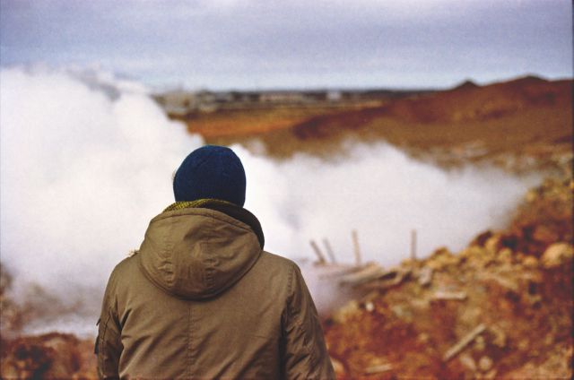 Individual wearing winter coat and beanie observing steaming geothermal area in rugged, remote location. The image captures the sense of exploration and adventure in a wild and solitary landscape. Ideal for use in travel blogs, adventure magazines, promotional material for outdoor gear, or websites related to geology and natural wonders.