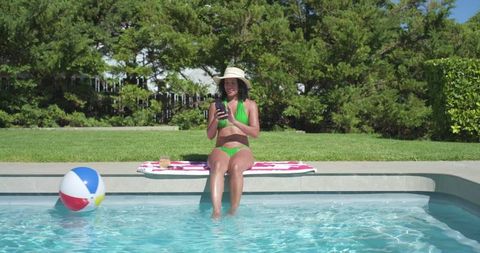 African american woman relaxing at backyard pool with phone, sun hat and colorful beach ball