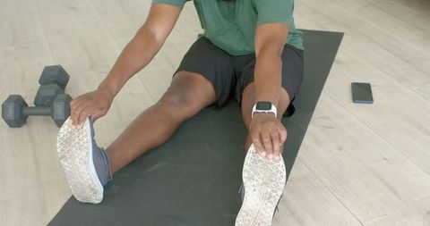 Man stretching hamstrings on mat at home wearing smartwatch with dumbbells