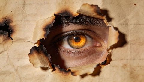 Amber eye peering through torn burnt parchment with charred edges close-up, golden iris