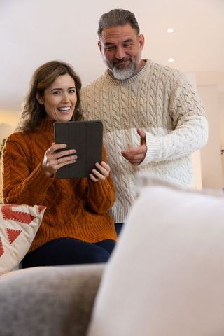 Middle-aged couple sharing tablet, smiling and enjoying cozy moment in knit sweaters