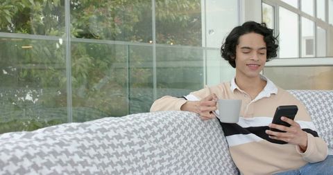 Young man relaxing on modern sofa scrolling smartphone and sipping coffee by window