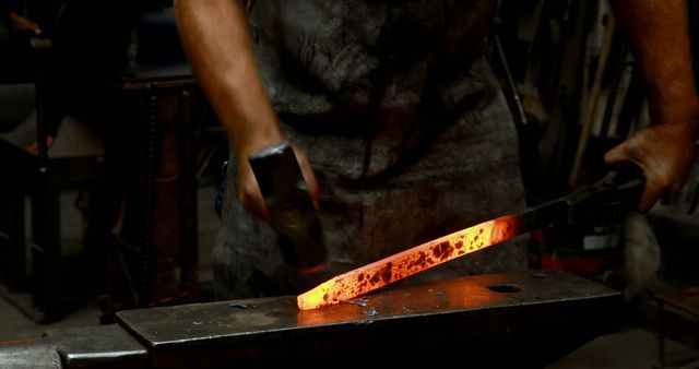 Blacksmith working on hot metal bar with hammer in dimly lit workshop. Artisan shapes glowing metal with precision and skill. Suitable for themes related to craftsmanship, traditional trades, hard work, artistry, industrial processes.