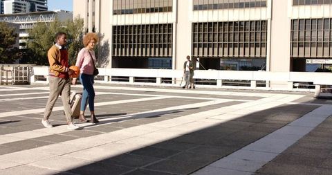 Diverse couple walking across rooftop pulling rolling suitcase carrying basketball