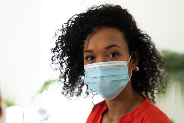 African American woman with curly hair wearing a facemask in an office setting. She is dressed in an orange blouse, emphasizing a professional and health-conscious environment. Ideal for use in articles or advertisements related to workplace safety, health protocols, pandemic measures, and professional settings during COVID-19.