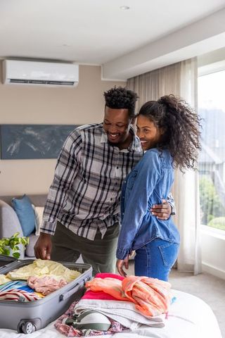 Happy diverse couple packing suitcase at home for weekend getaway, sharing warm moment