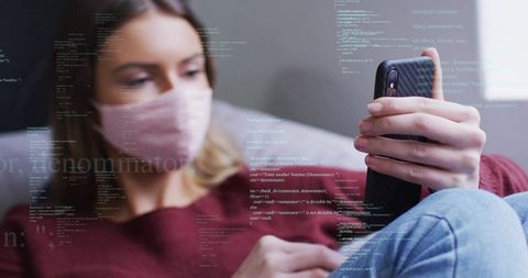 Masked woman scrolling smartphone with coding overlays reclining on couch in soft natural light