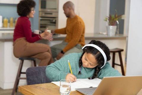 Multiracial family child doing homework with headphones while parents chatting over coffee