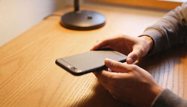 Hands holding smartphone in landscape orientation on wooden desk with warm lamp light