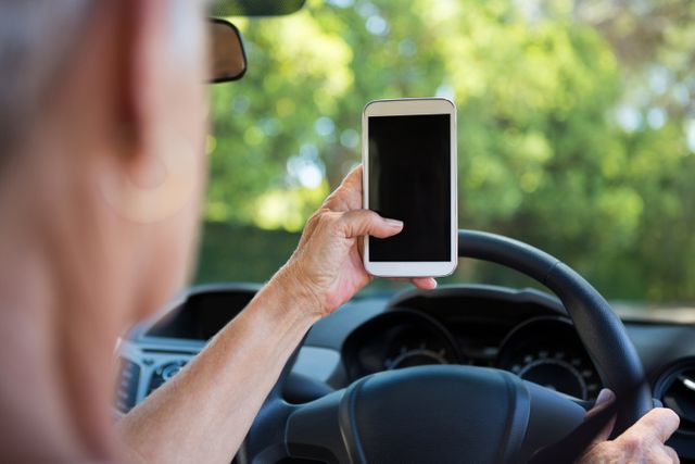 Senior woman holding a mobile phone while driving a car. This image can be used in campaigns promoting road safety, highlighting the dangers of distracted driving, or illustrating the use of technology by elderly people. It is suitable for articles, advertisements, and educational materials on safe driving practices and mobile phone usage.