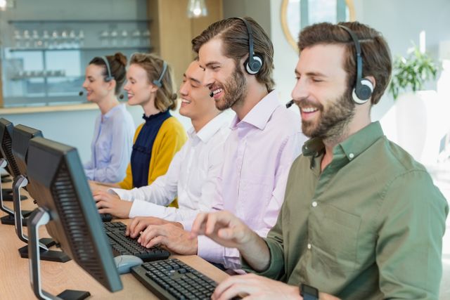 Group of diverse customer service executives working at computers, wearing headsets, and smiling. They are seated in a modern office environment, cooperating to provide support. Perfect for business, communication, teamwork, and customer care concepts.