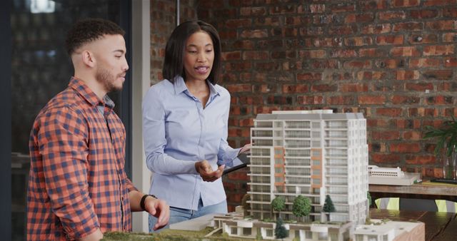 Two architects involved in a discussion about a scale model of a modern building in an office. This image can be used for illustrating teamwork in architecture, business meetings, or office settings, and is ideal for materials relating to architectural planning, collaborative work environments, and construction projects.