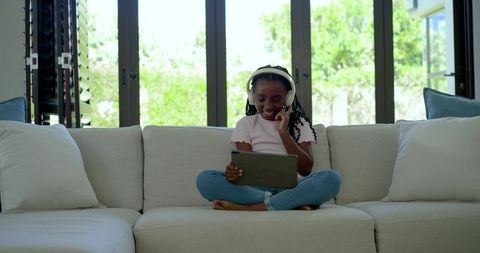 African american girl using tablet with headphones on cozy sofa for remote learning