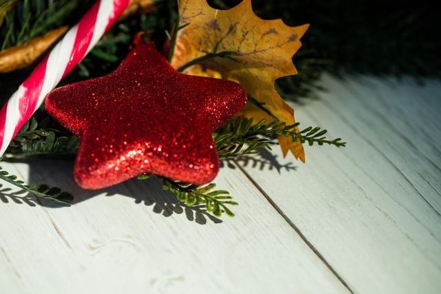 Close-up of red star-shaped Christmas ornament and candy cane placed on pine leaves on a white wooden plank. Ideal for holiday greeting cards, festive season advertisements, and social media posts celebrating Christmas.