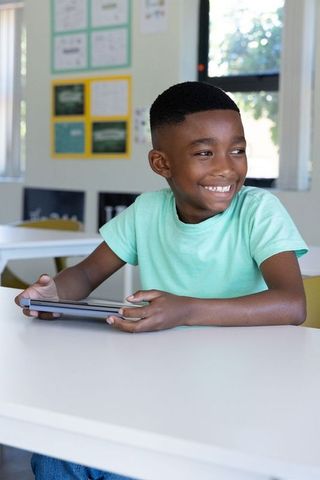 African american boy smiling using tablet in bright classroom for digital learning