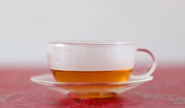 Clear glass cup filled with hot tea, resting on a matching glass saucer on a red tablecloth. Light steam is visible, indicating the warmth of the tea. Ideal for concepts related to relaxation, tea time, morning routine, health benefits of tea, and keeping warm. Suitable for articles, blog posts, advertising for tea brands, wellness and lifestyle campaigns, or kitchen decor showcases.