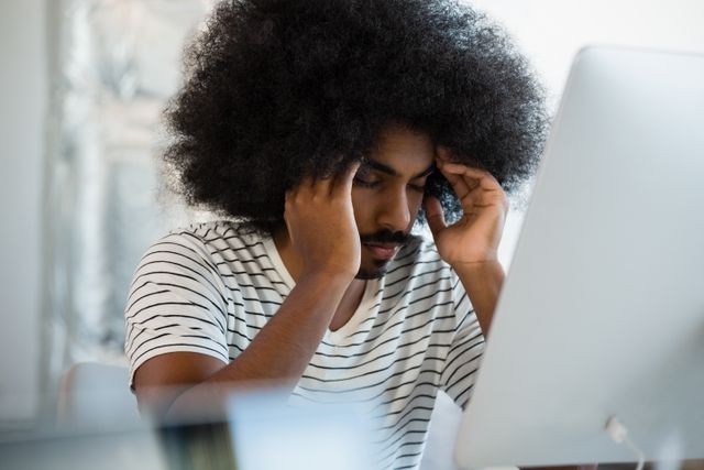 Young man with afro hair sitting at desk in creative office, eyes closed, holding head in hands. Ideal for illustrating workplace stress, mental health awareness, fatigue, and the challenges of modern work environments.