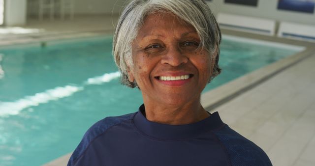 Senior woman standing by indoor swimming pool, smiling and enjoying moment. Perfect for illustrating vibrant senior living, wellness campaigns, active aging, and promotional material for fitness centers or aquatic programs dedicated to elderly individuals.