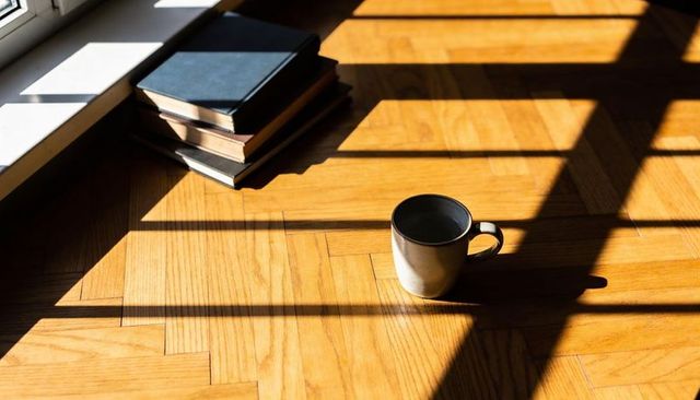 Sunlit ceramic mug casting long shadow on parquet floor with stack of books