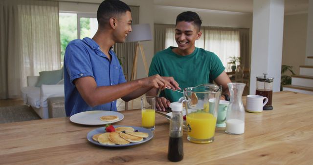 Happy biracial gay male couple having pancakes for breakfast and laughing in kitchen. staying at home in isolation during quarantine lockdown.