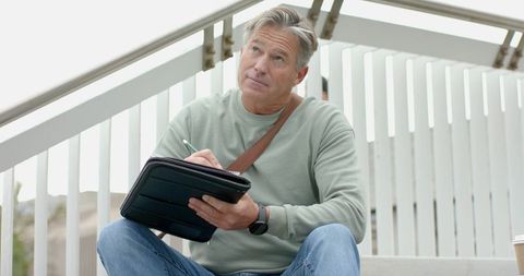 Mature man writing in tablet folio on white steps, contemplating ideas for work or journaling