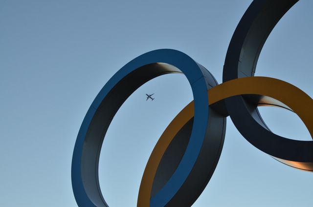Airplane flying through city sculpture with large interlocking rings dominating foreground against clear blue sky. This can symbolize travel, global connectivity, modern architecture, and aviation. Suitable for use in designs related to travel industry, aviation, contemporary art displays, or modern architectural projects.