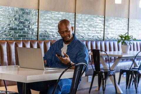 African man working remotely in cafe using laptop and phone, modern coworking workspace