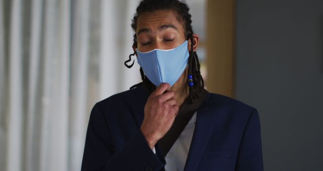 Businessman wearing face mask indoors touching his chin. Suitable for themes on workplace health precautions, professional life during the pandemic, health and safety at work, contemplative business moments, and maintaining hygiene in an office environment.