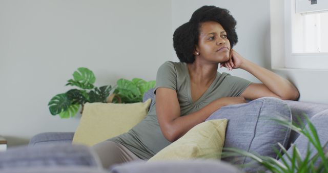 The image shows an African American woman sitting on a couch in a modern living room. She is in a thoughtful or contemplative mood, leaning on her hand with a peaceful expression. This photo can be used in lifestyle blogs, articles on mental health and wellbeing, or settings depicting relaxation and comfort at home.