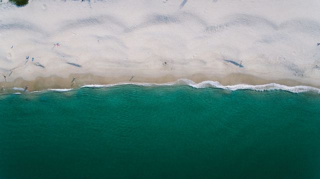 Aerial photograph showcasing a tranquil sandy beach with gentle waves washing ashore and emerald green water. Ideal for travel and tourism websites, beach destinations promotions, nature conservation materials, summer vacation ideas, and relaxation-themed content.