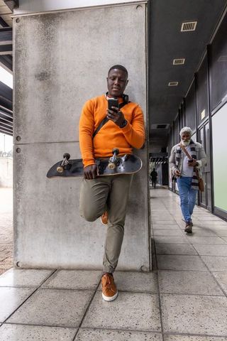 Urban skateboarder leaning on concrete wall checking smartphone in orange sweatshirt